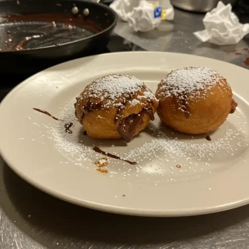 Two deep-fried s'mores dusted with powdered sugar on a chipped plate, surrounded by kitchen clutter.