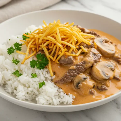 Close-up top-down view of the finished Brazilian Beef Stroganoff in a white bowl over rice, garnished with Batata Palha (shoestring potato sticks) and fresh parsley.