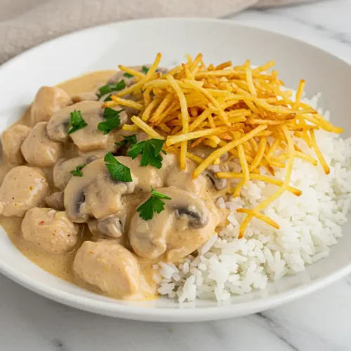 Close-up top-down view of the finished Brazilian Chicken Stroganoff served over white rice in a bowl, garnished with shoestring potatoes and fresh parsley.