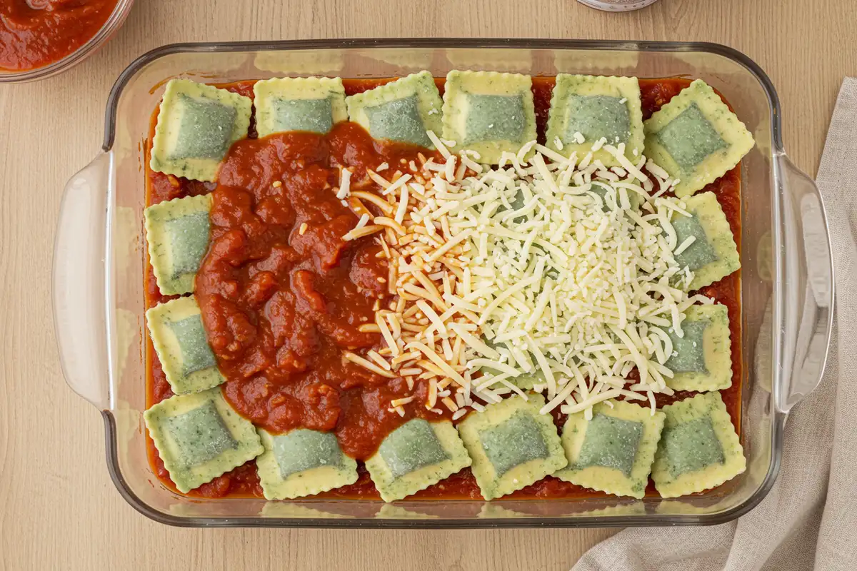 Close-up top-down view of Baked Spinach Ravioli casserole being layered in a 9x13 dish: frozen ravioli covered with marinara sauce and sprinkled with mozzarella and Parmesan, natural home-assembly feel.