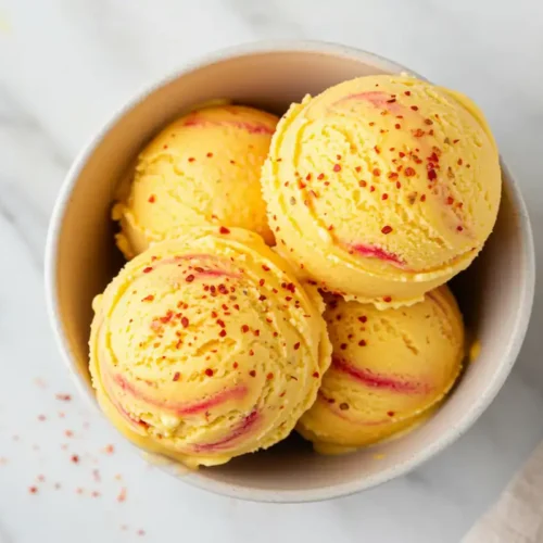 Extreme close-up overhead view of creamy mango ice cream scoops in a bowl, showing distinct Tajin swirls within and a fresh sprinkle of Tajin seasoning on top, on a light marble surface.