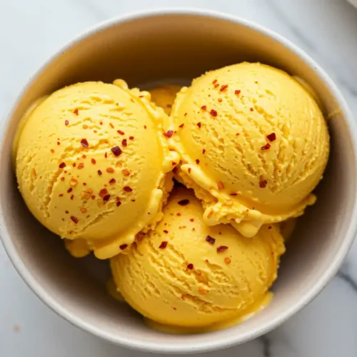 Extreme close-up overhead view of creamy, vibrant orange spicy mango ice cream scoops in a bowl, showing visible reddish spice specks throughout and a chili lime seasoning garnish on top.