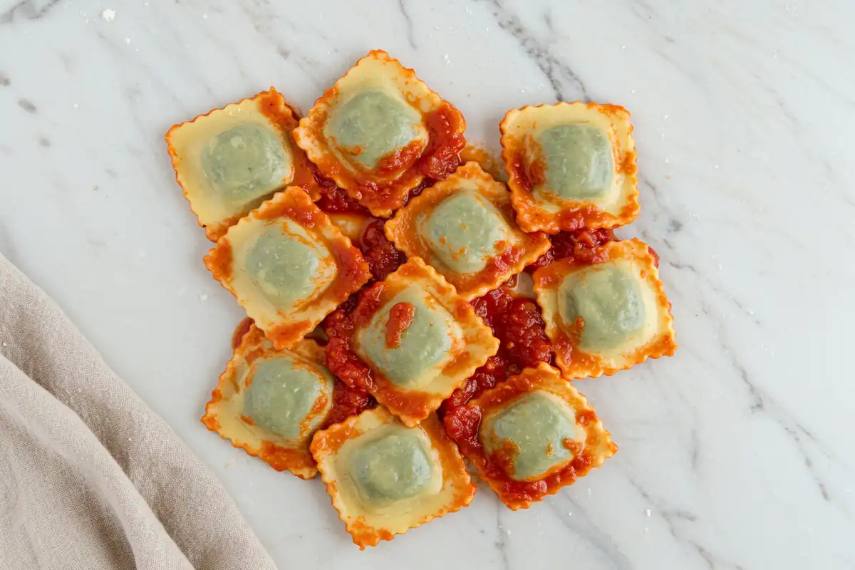 Overhead close-up of spinach and ricotta ravioli tossed in a bright simple marinara sauce, presented on a light marble surface with a beige cloth, captured in soft natural light for a realistic feel.