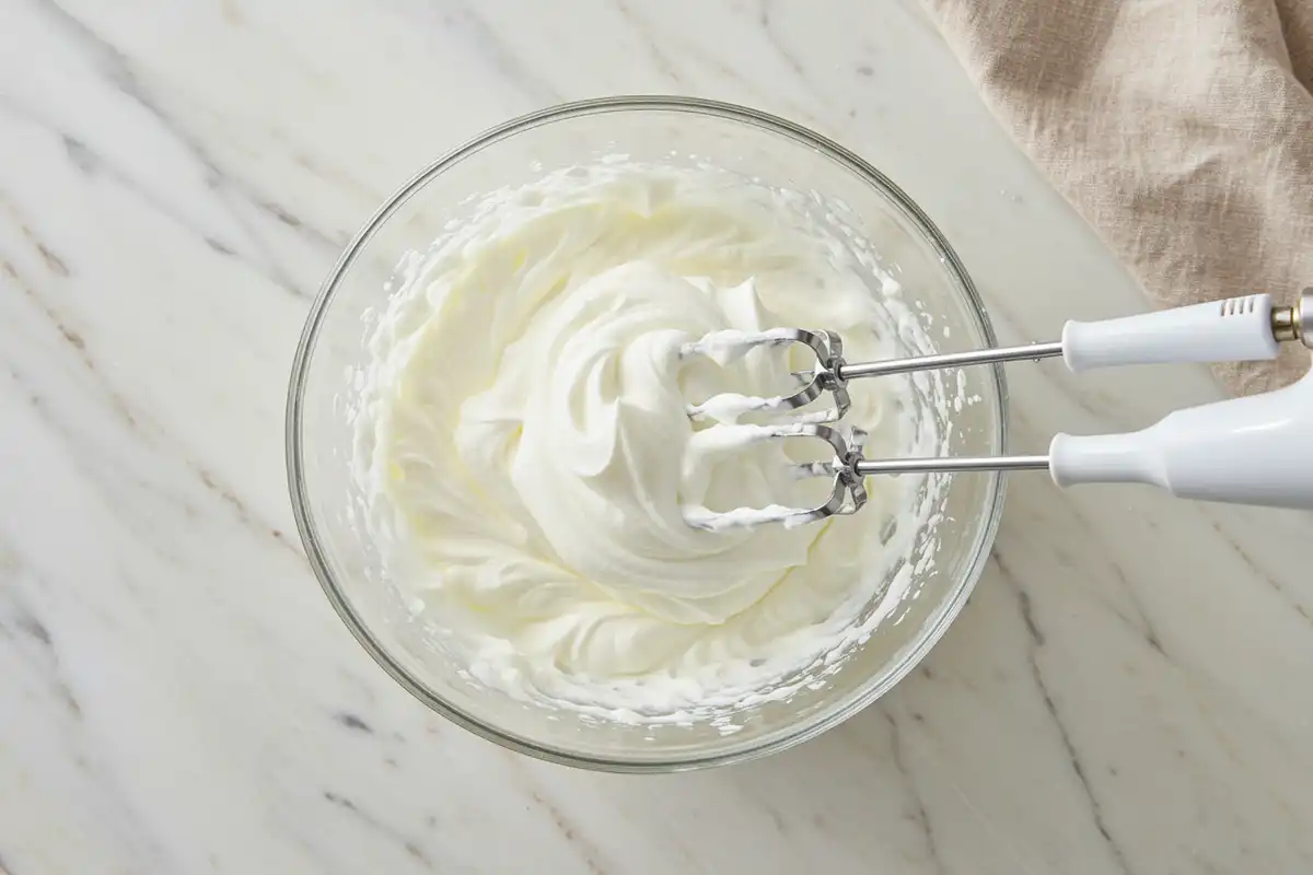 Close-up top-down view of Greek yogurt being whipped with hand mixer beaters in a bowl with subtle wear, showing the yogurt becoming light, airy, and forming soft, billowy peaks, with a natural, home-cooked feel.